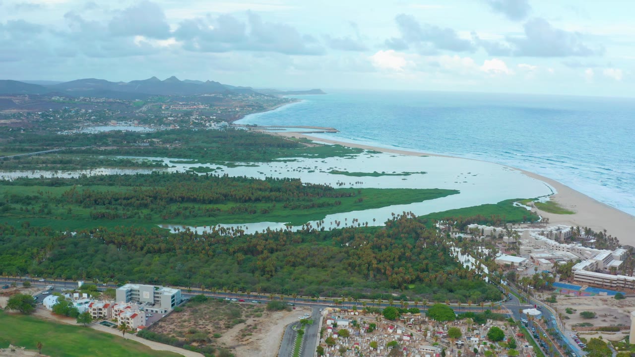 Drone view of lagoon, beach, coastal buildings, roads, ocean, and cloudy sky.