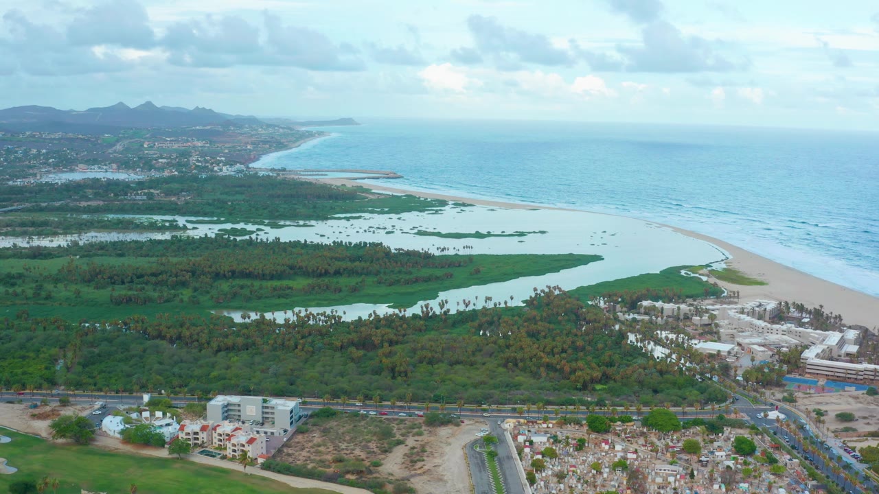 Aerial view in San Jose Del Cabo with the lagoon and green wetland centered below the beach curve, ocean to the right, buildings and roads along the lower edge, and no visible people.