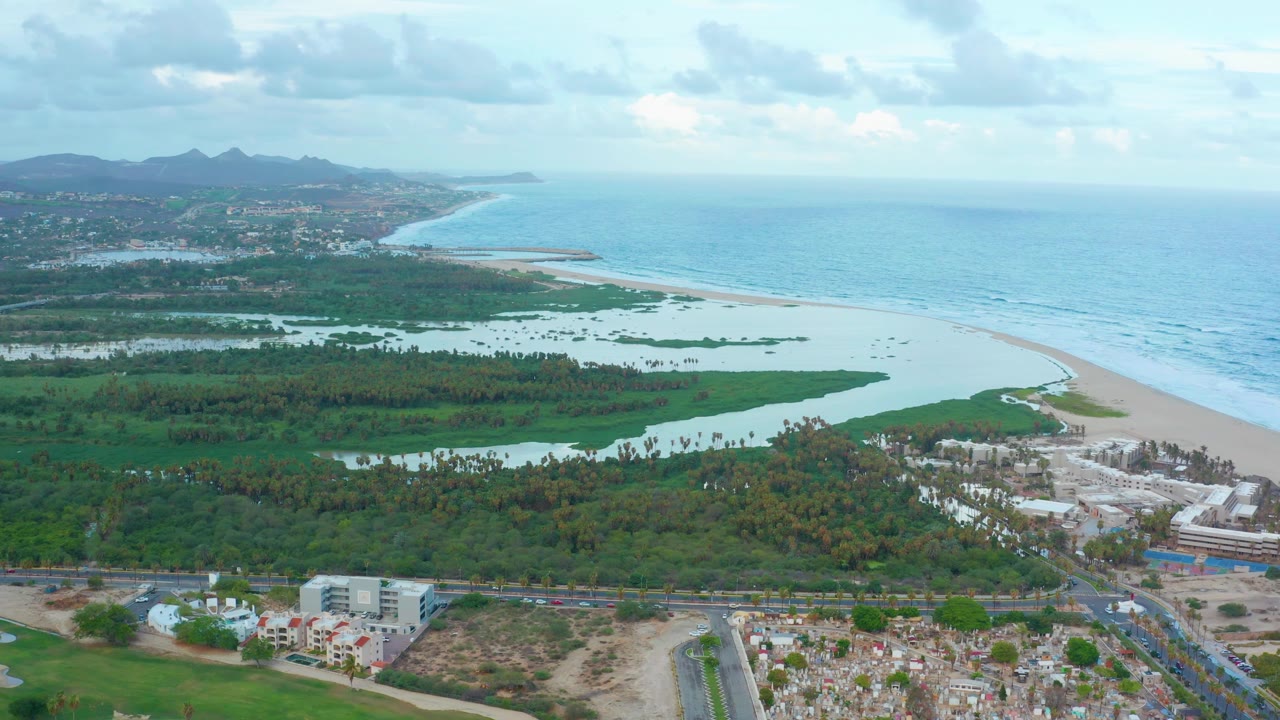 Wide San Jose Del Cabo aerial with green lagoon water and vegetation in the midground, sandy beach and blue ocean beyond, roadside development below, distant hills at left, and no visible people.