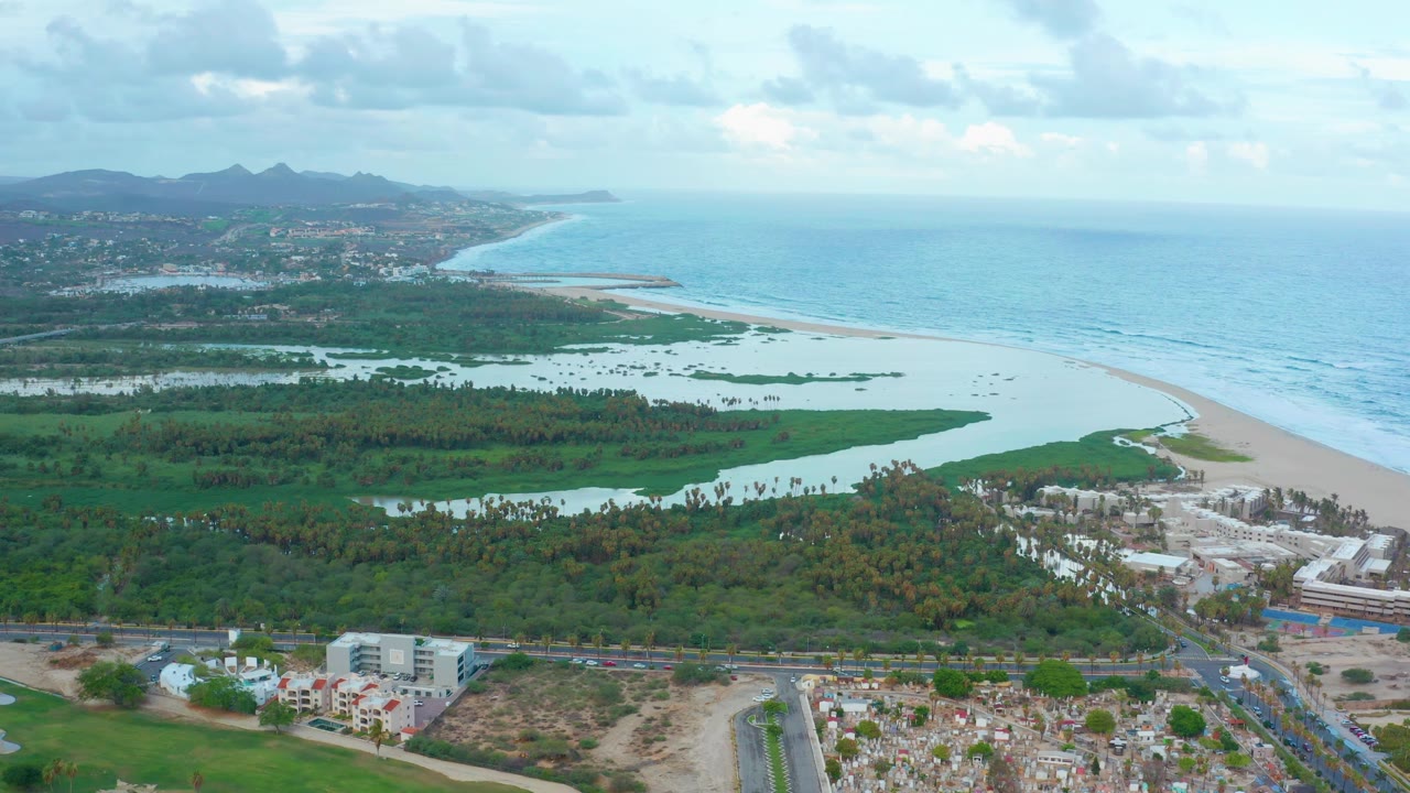 San Jose Del Cabo aerial frame with the lagoon spread across the center, the ocean horizon to the right, a pale beach between them, roads and low buildings in the foreground, and no visible people.