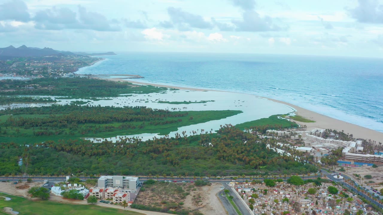 High aerial frame in San Jose Del Cabo with lagoon channels across green vegetation, a long sandy beach beside blue ocean, foreground development, hazy hills at left, clouded sky, and no visible people.