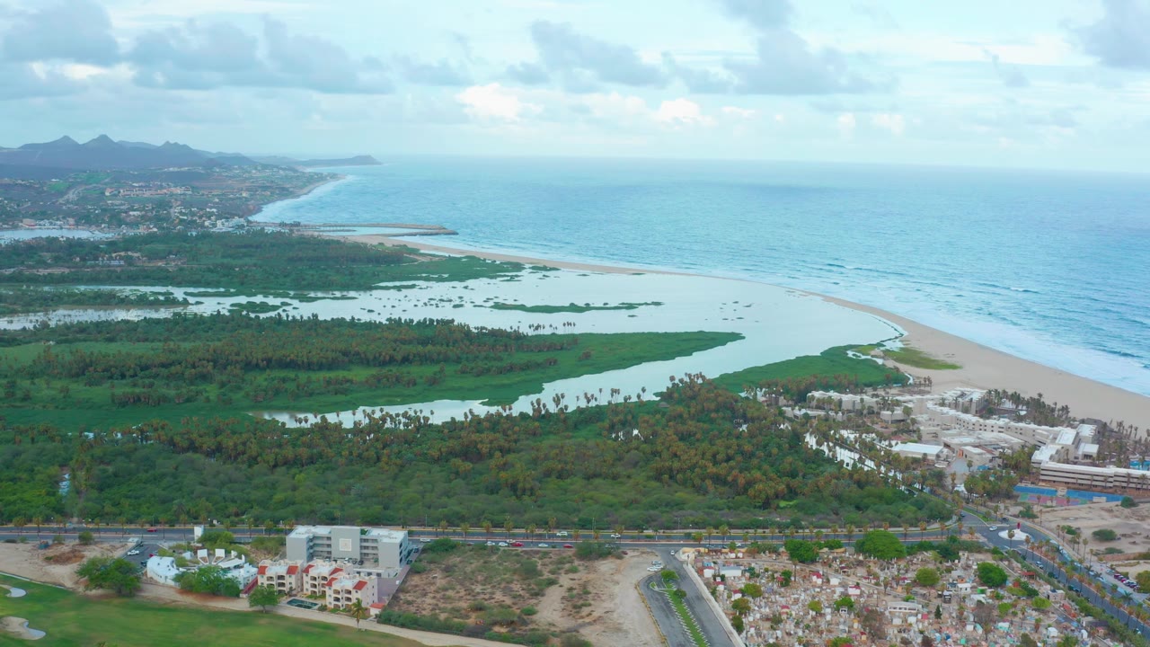 Aerial coastal landscape in San Jose Del Cabo showing a bright ocean horizon, sandy shoreline, lagoon water cutting through green land, foreground roads and low buildings, distant hills, and no visible people.