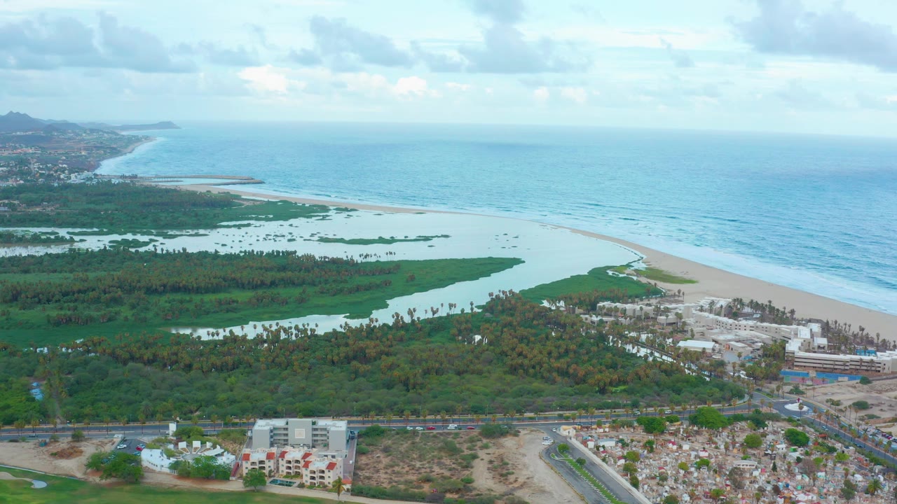 Aerial view over San Jose Del Cabo showing a coastal lagoon beside a pale beach, turquoise ocean, inland greenery, roads, low buildings, cloudy sky, and no visible people.
