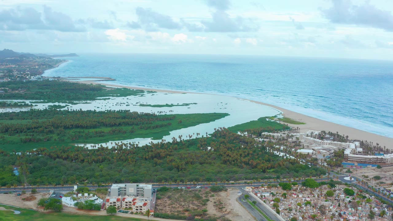 San Jose Del Cabo aerial frame with the lagoon and beach centered, ocean stretching across the right side, green wetland areas inland, buildings and roads below, and no visible people.