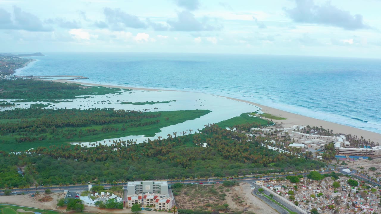 A wide aerial frame over San Jose del Cabo shows green wetlands and pale lagoon water before a sandy beach and blue ocean, with roads and buildings in the foreground and no visible people.