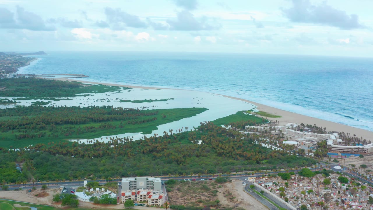 A high aerial view of San Jose del Cabo shows the lagoon and wetlands stretching across the middle of the frame, the beach running diagonally toward the ocean, and foreground structures below, with no visible people.