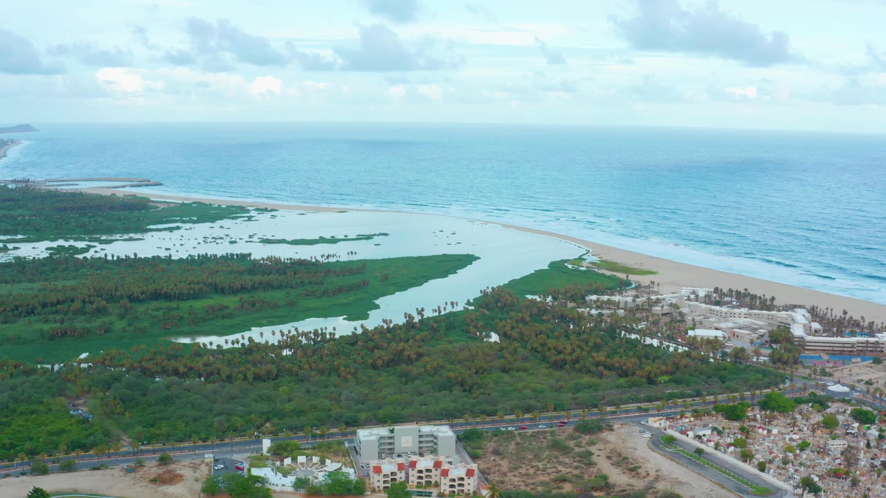 Wide aerial view over San Jose Del Cabo showing a green lagoon, pale beach, blue ocean, coastal buildings, a road, and no visible people at the start of the clip.