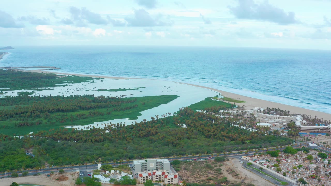 Aerial frame from San Jose Del Cabo with lagoon water and greenery across the middle, surf lines on the beach, low buildings near the road, and no visible people.