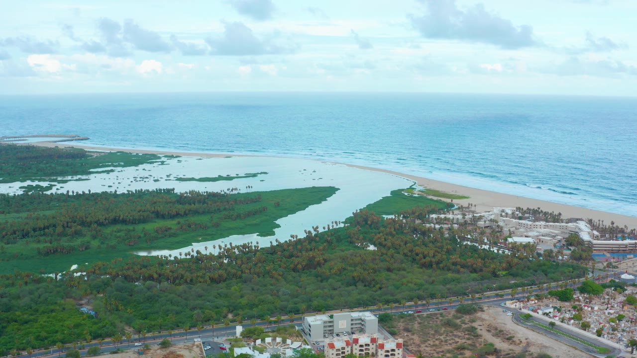 Second aerial frame of San Jose Del Cabo coastline showing the lagoon wrapping around dense vegetation, surf along the beach, distant ocean horizon, buildings, road, and no visible people.
