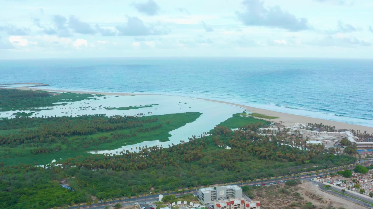 A high aerial view over San Jose del Cabo shows a green lagoon landscape, pale beach, blue ocean horizon, shoreline buildings, a road, and no visible people.