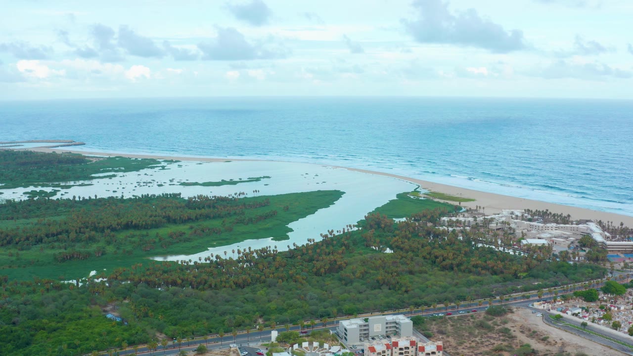 A broad San Jose del Cabo aerial frame shows the ocean horizon across the top, a light sand beach, lagoon channels in green vegetation, low buildings, roadways, and no visible people.