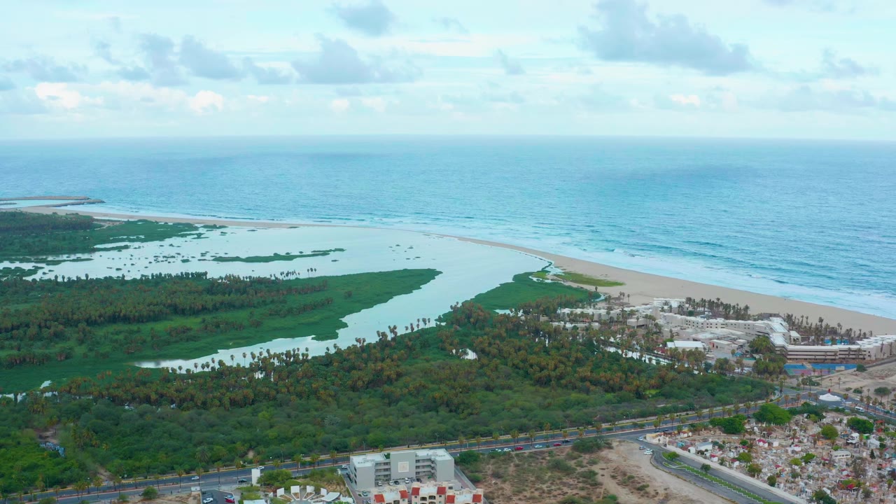 San Jose del Cabo aerial frame with the lagoon stretching across the left side, beach and surf running along the coast, greenery below, and built development near the lower right, with no visible people.