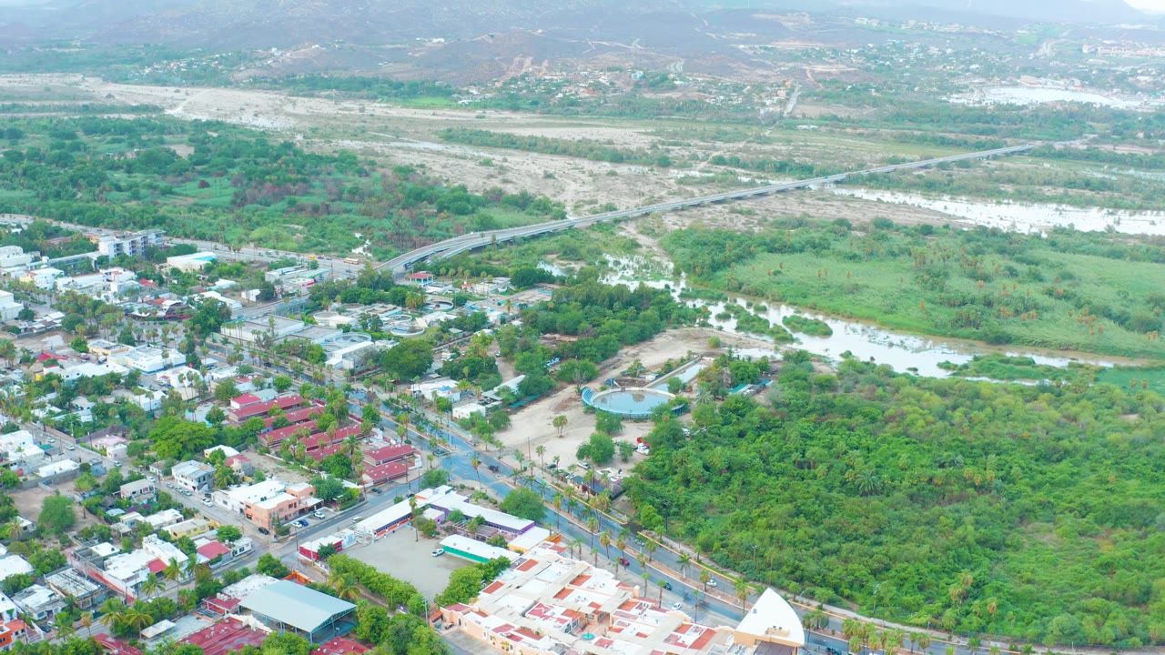 A drone-height view over San Jose del Cabo showing town blocks at the lower edge, a wide green river corridor, a diagonal bridge, pale riverbed terrain, distant hills, and no visible people.