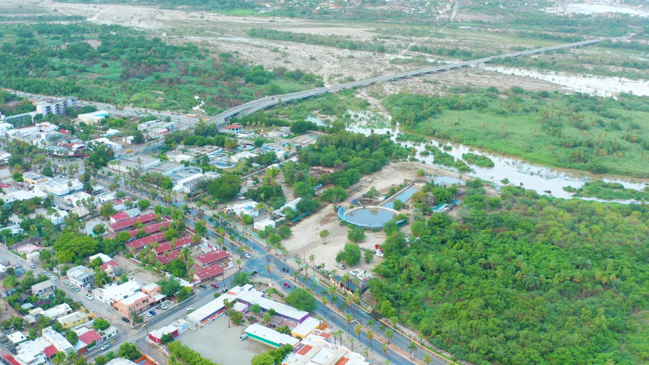 Aerial frame over San Jose Del Cabo shows pale floodwater crossing a river corridor near roads, low buildings, red rooftops, dense green trees, and no visible people.