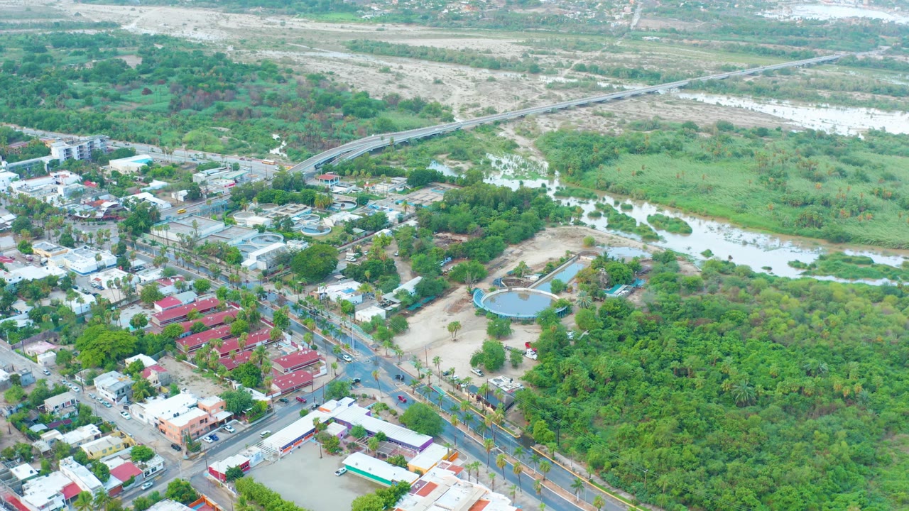 Aerial view in San Jose Del Cabo shows a green river channel with muddy water, nearby streets, compact buildings, a wooded area, and no visible people.