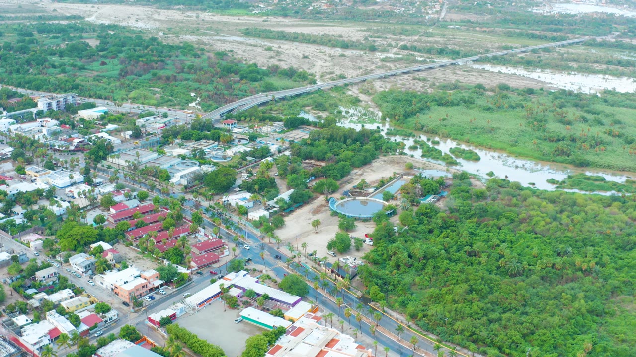 High aerial frame over San Jose Del Cabo shows roads and rooftops in the foreground, a broad tree canopy in the center, floodwater through the riverbed, and no visible people.