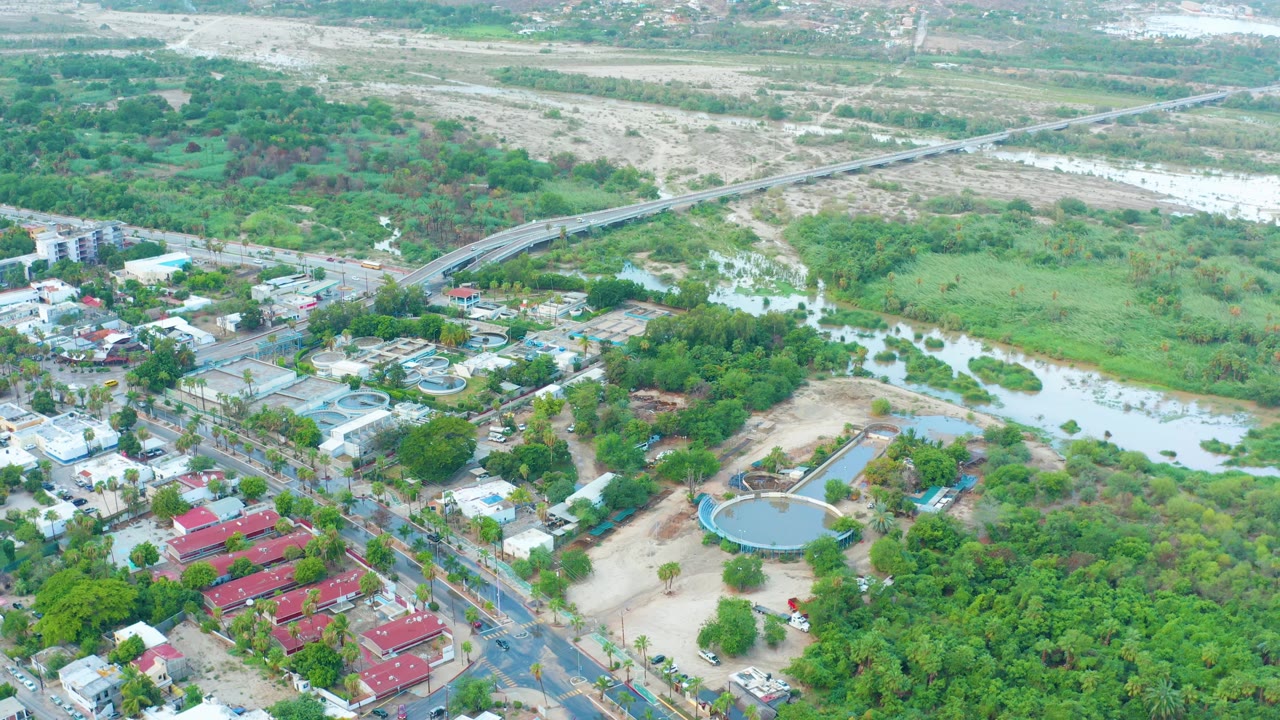 Opening aerial frame over San Jose Del Cabo, Mexico, showing floodwater running through a green river corridor near roads, homes, red-roofed buildings, and a distant bridge, with no visible people.