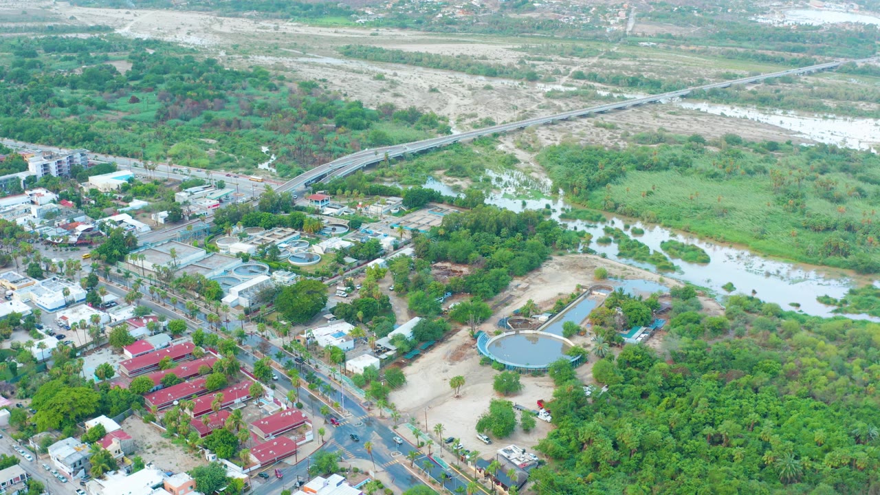 San Jose Del Cabo aerial view showing the swollen river and wet low-lying land between a residential road grid, red-roofed buildings, dense green trees, and the bridge beyond, with no visible people.