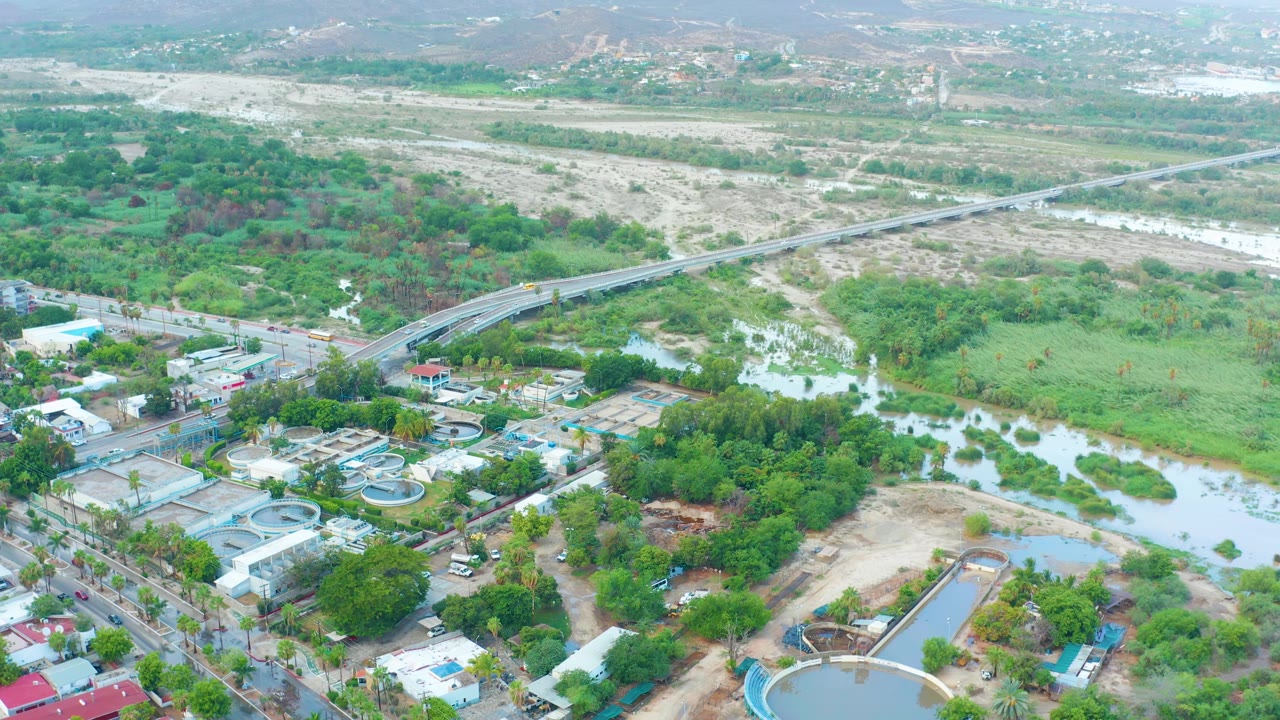 High aerial view over San Jose del Cabo showing the river channel, road bridge, low buildings, circular pools, green vegetation, hazy hills, and no visible people at the start of the clip.