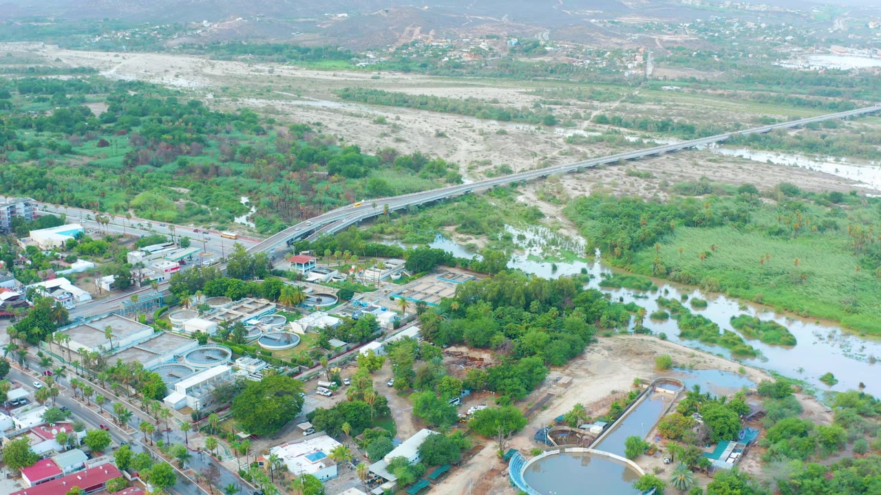 Mid-clip aerial over San Jose del Cabo with a broad pale river channel, long bridge, roadside buildings, circular pools near the lower edge, dense green patches, and no visible people.