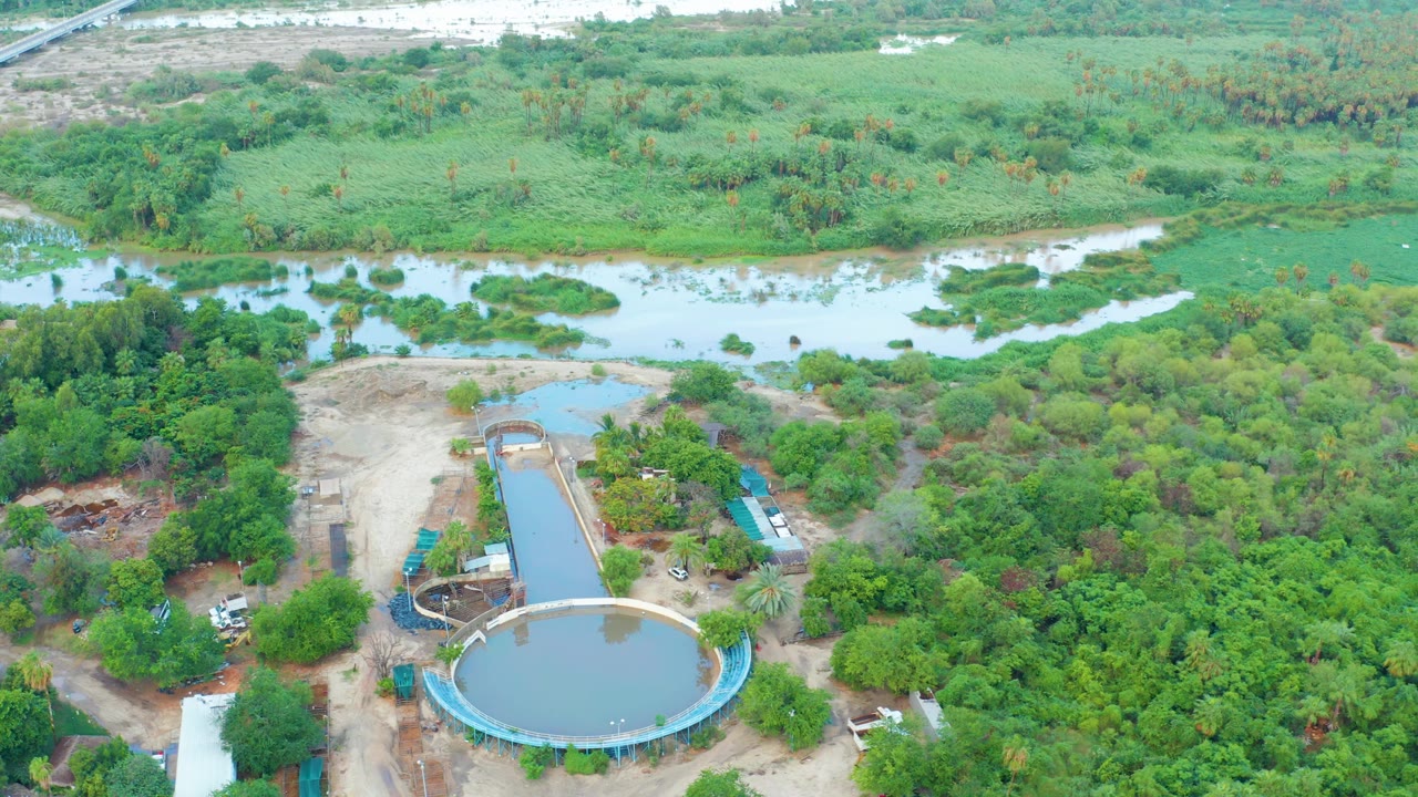 Opening aerial frame in San Jose Del Cabo shows a circular tank, rectangular basin, dirt service area, river channel, dense green trees, and no visible people.