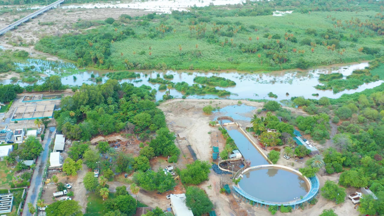 Overhead frame from San Jose del Cabo showing a pale river behind dense green vegetation, a circular water treatment tank, dirt lots, roads, and no visible people.