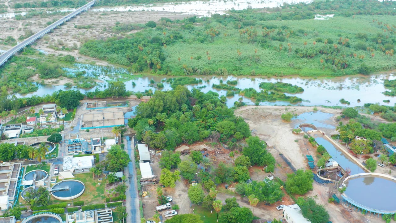 Drone-like view over San Jose del Cabo with a large circular tank at lower right, smaller tanks at left, dense trees in the center, river pools beyond, and no visible people.