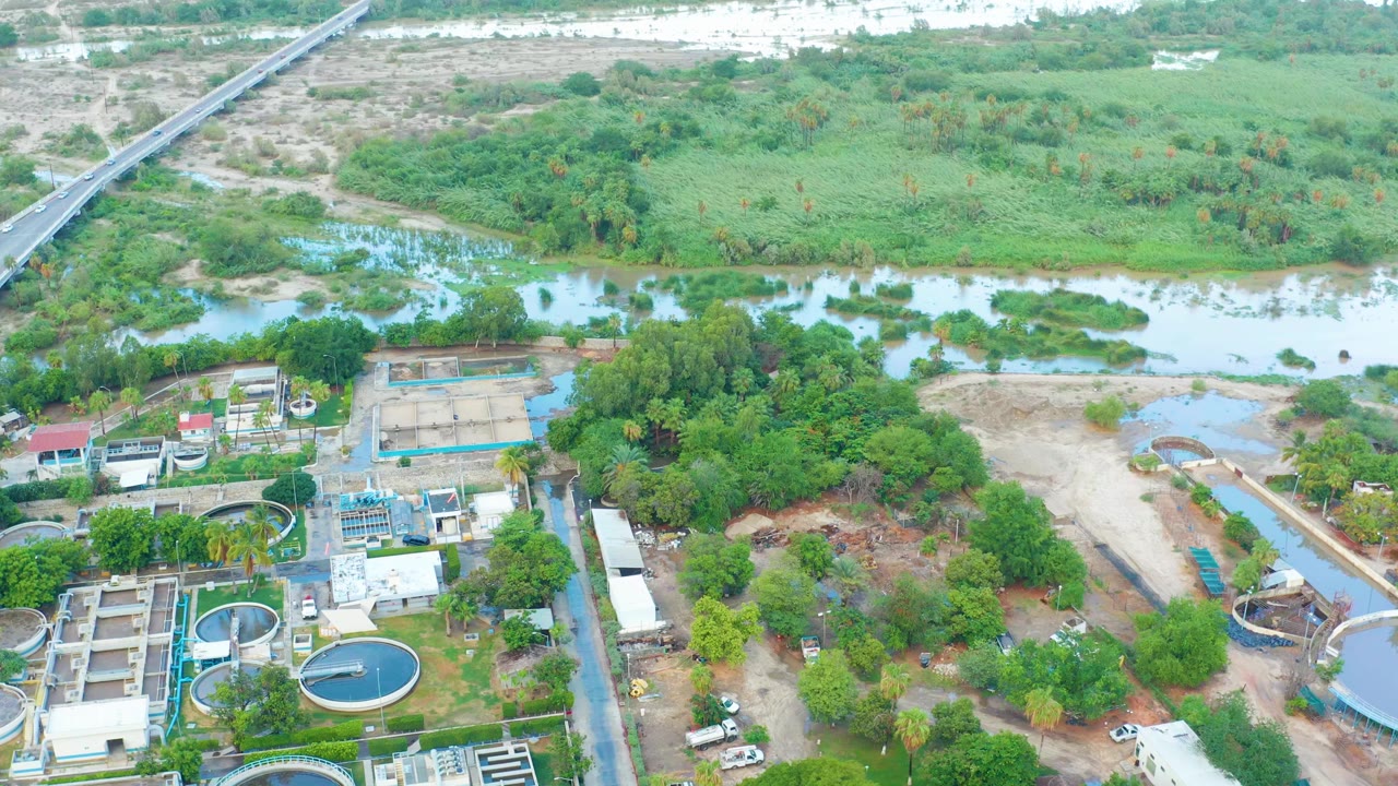 Second aerial frame in San Jose del Cabo showing the plant's round tanks and rectangular concrete treatment areas in the lower frame, with river water and thick green vegetation behind them and no visible people.