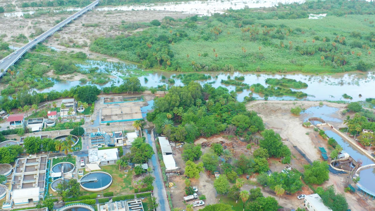 Mid-clip aerial frame over San Jose del Cabo's water treatment facility, with circular tanks in the lower left, a curved channel at right, a tree-covered riverbank, broad green vegetation, and no visible people.