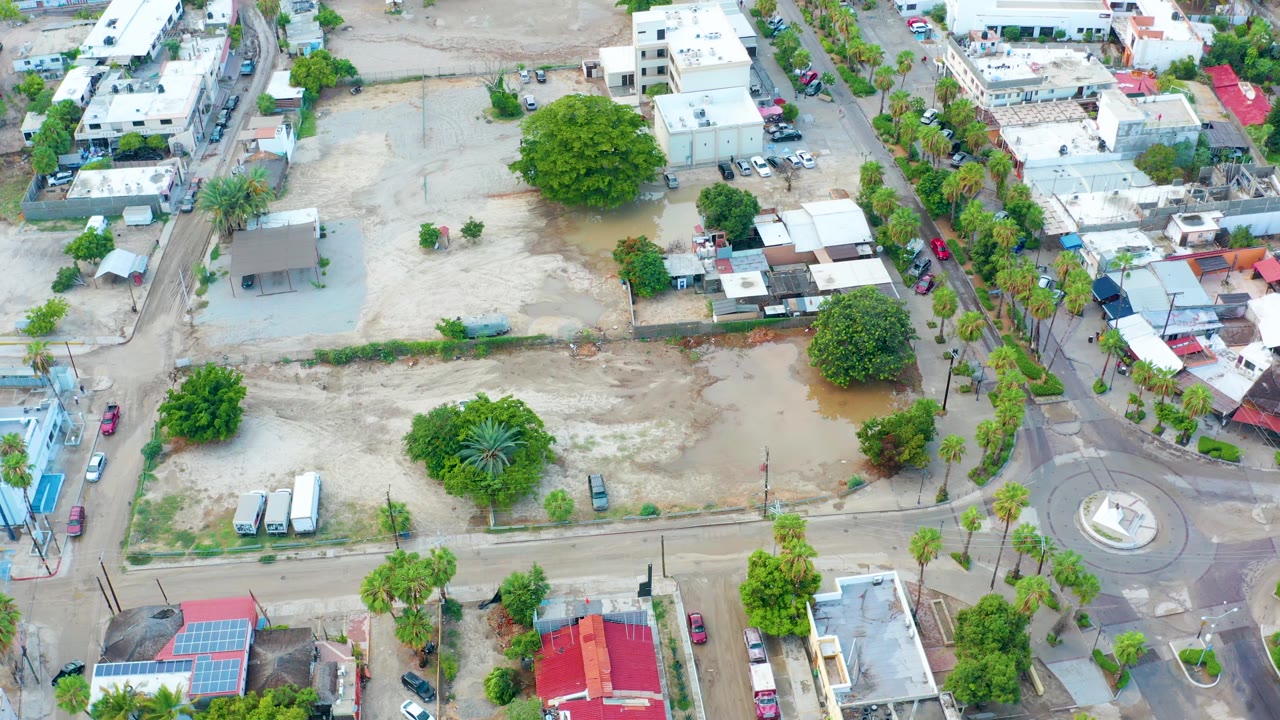 High aerial view in San Jose Del Cabo, Mexico, showing a sandy urban lot with shallow brown floodwater, a roundabout at lower right, low buildings, trees, parked vehicles, and no visible people.
