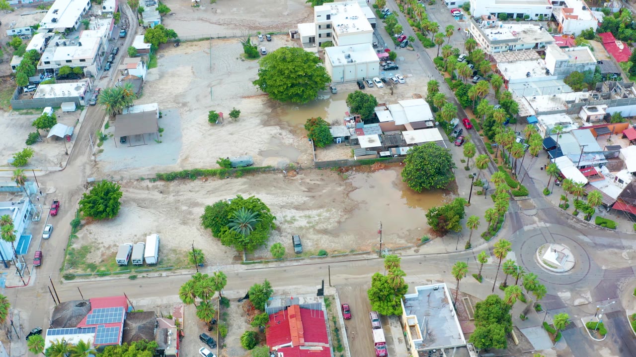 Aerial frame over San Jose Del Cabo with floodwater pooled through the center of a vacant sandy lot, palm-lined streets, nearby low-rise buildings, parked cars, and no visible people.