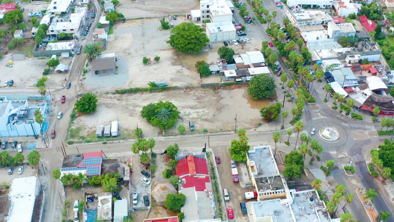 A second aerial view of the San Jose del Cabo lot shows brown pooled water near a large tree, with low buildings, parked vehicles, and no visible people.
