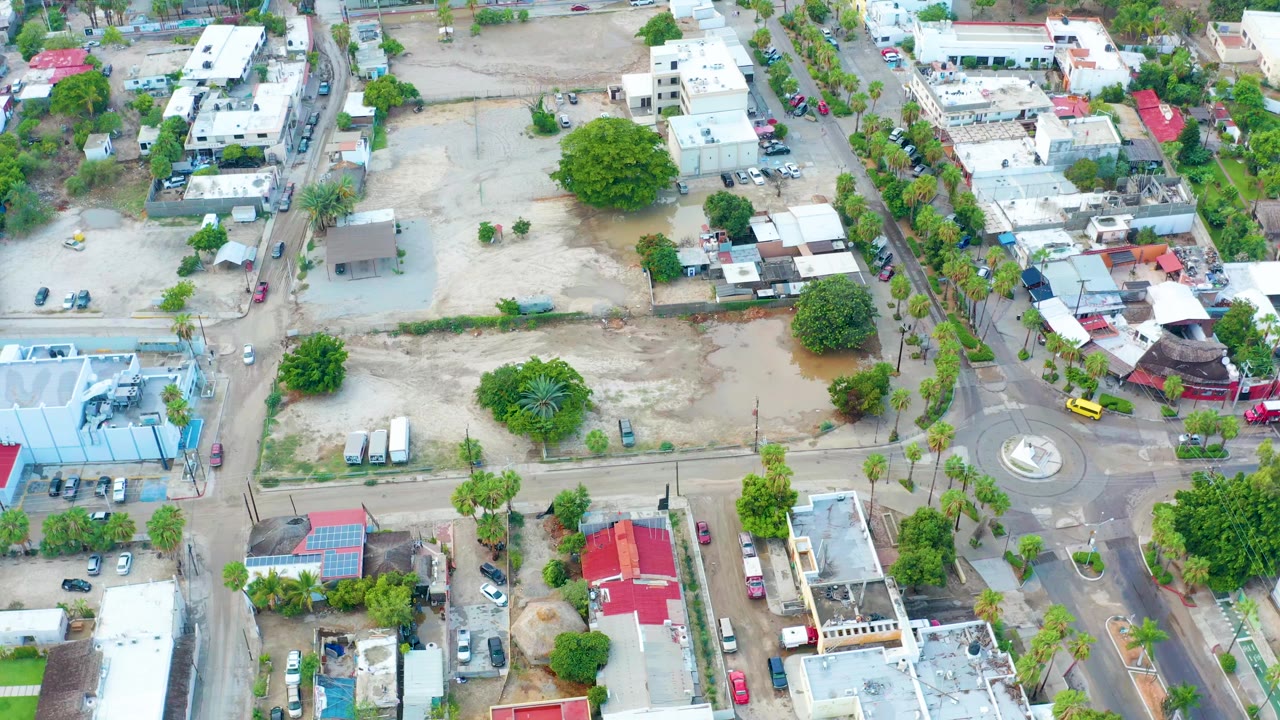 Aerial neighborhood view with diagonal roads, parked vehicles, trees, and open lots.