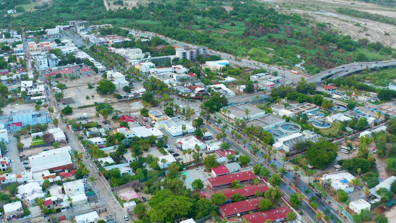 Aerial frame in San Jose del Cabo showing residential blocks, palm-lined streets, circular blue water basins, parked vehicles, and green terrain in the background, with no visible people.