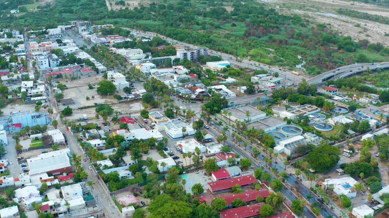 San Jose del Cabo aerial view with a road overpass at the right edge, white homes, red roofs, tree cover, and a green hillside beyond the developed blocks, with no visible people.