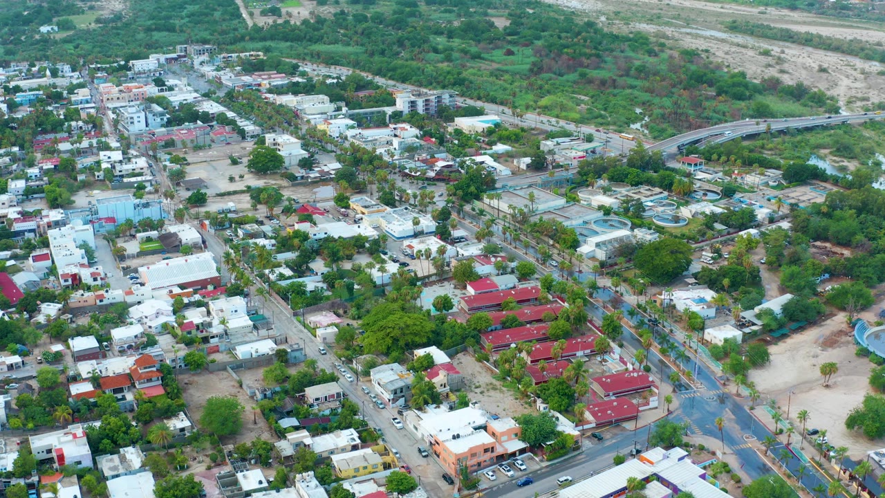 Drone view over low-rise houses, green trees, crossing streets, and open land beyond town.