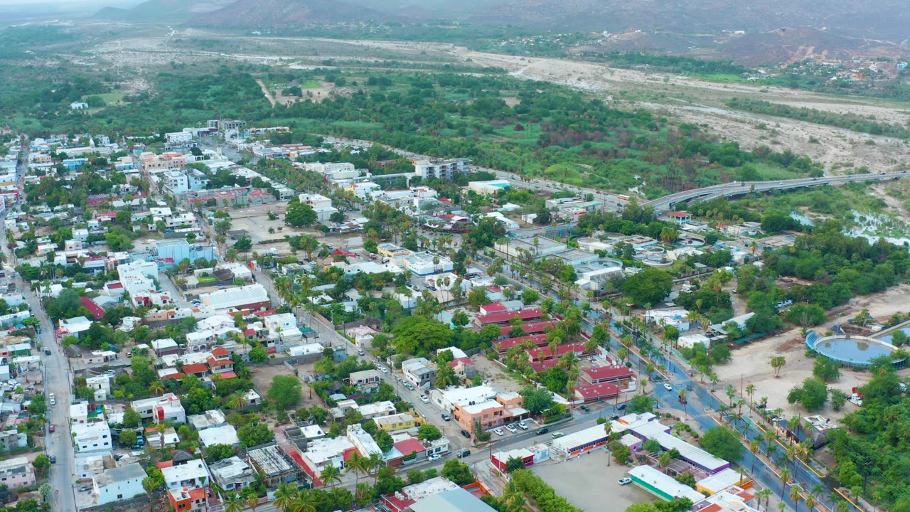 Opening aerial frame over San Jose del Cabo showing low-rise buildings, tree-lined streets, sandy lots, a dry riverbed beyond the town, hazy hills in the distance, and no visible people.