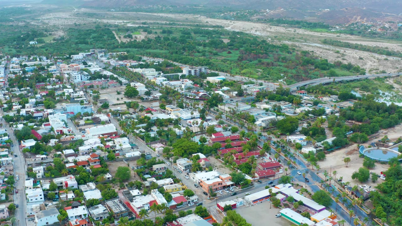 Third frame shows San Jose del Cabo from above with a broad town block in the lower frame, a tree-filled midground, a pale dry riverbed crossing the background, and no visible people.