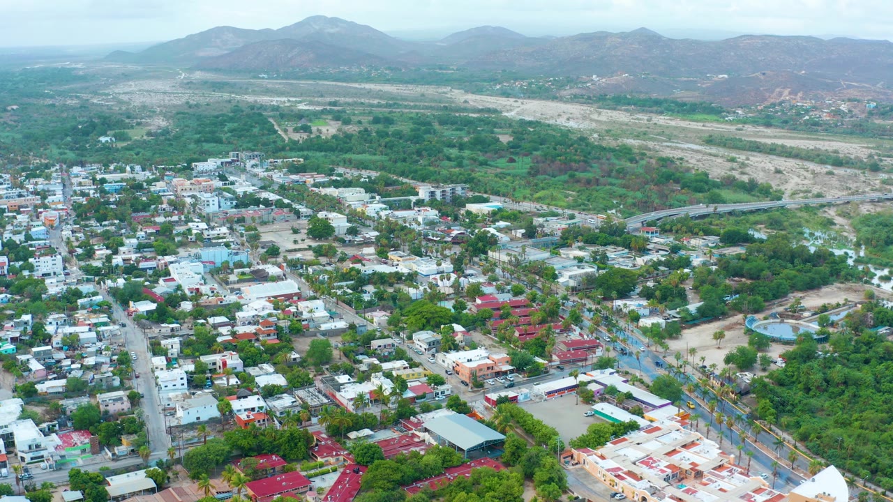 Wide drone view across rooftops, roads, trees, dry riverbed, and mountains.