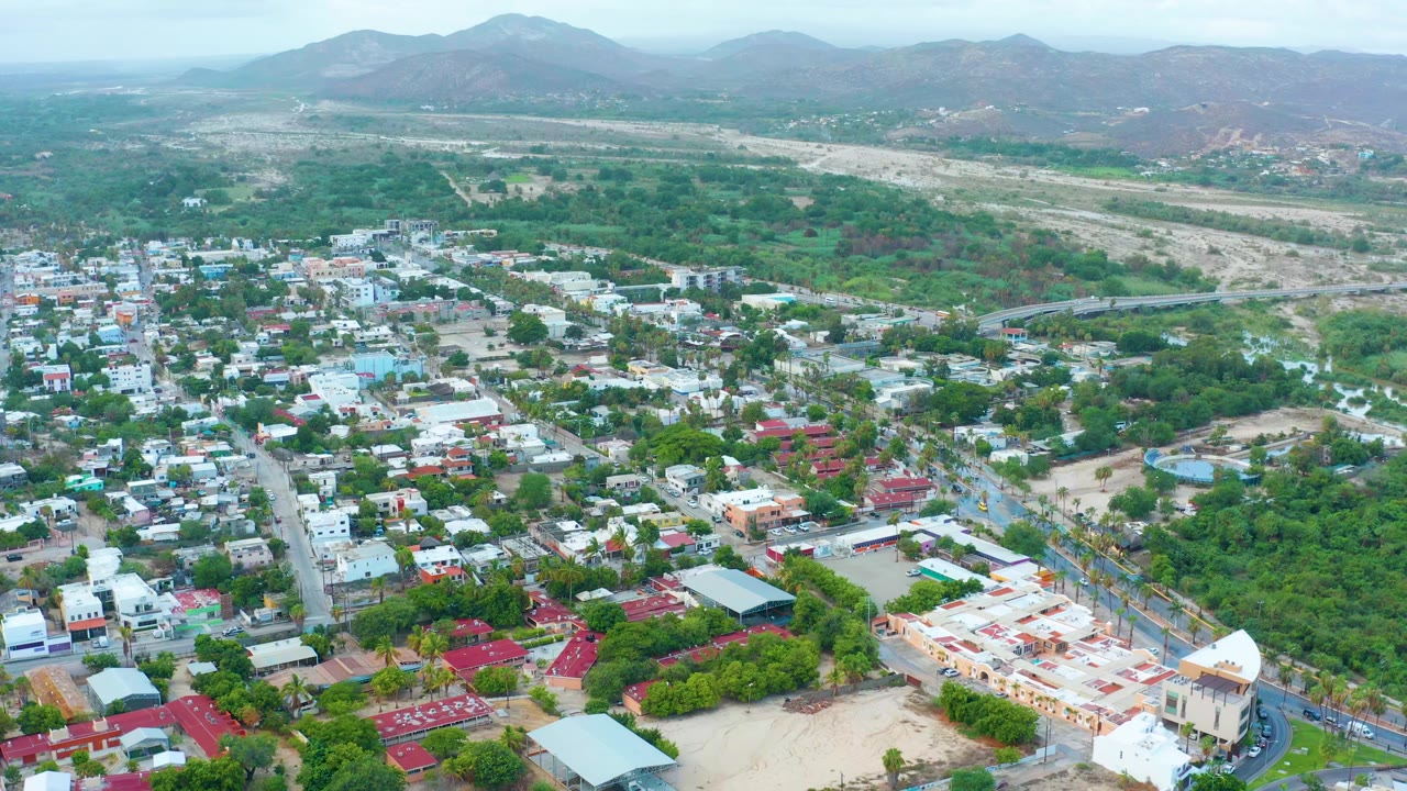 A second elevated view of San Jose Del Cabo keeps the residential blocks and tree canopy in the lower frame, with the pale wash and hazy mountain ridge stretching behind them and no visible people.