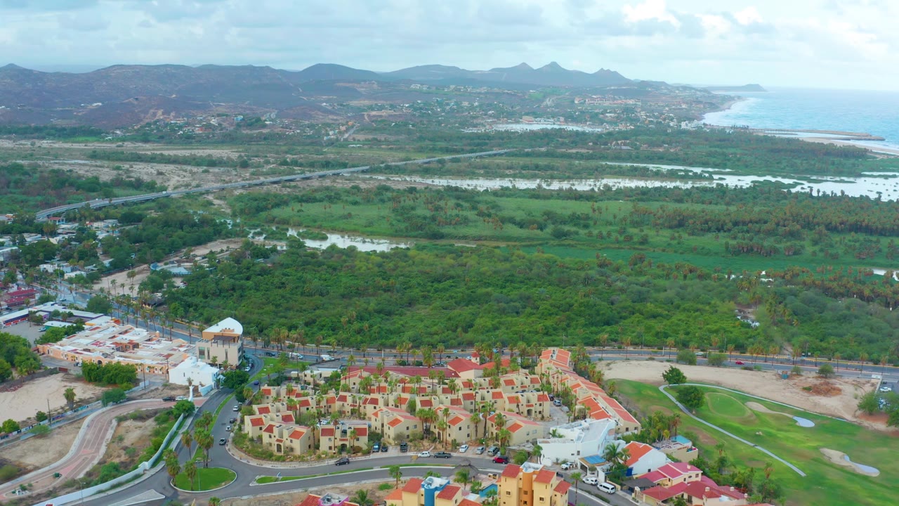 Aerial view in San Jose del Cabo with the red-roofed neighborhood lower in frame, curved roads at left, green lowlands and reflective water in the center, hills on the horizon, and no visible people.