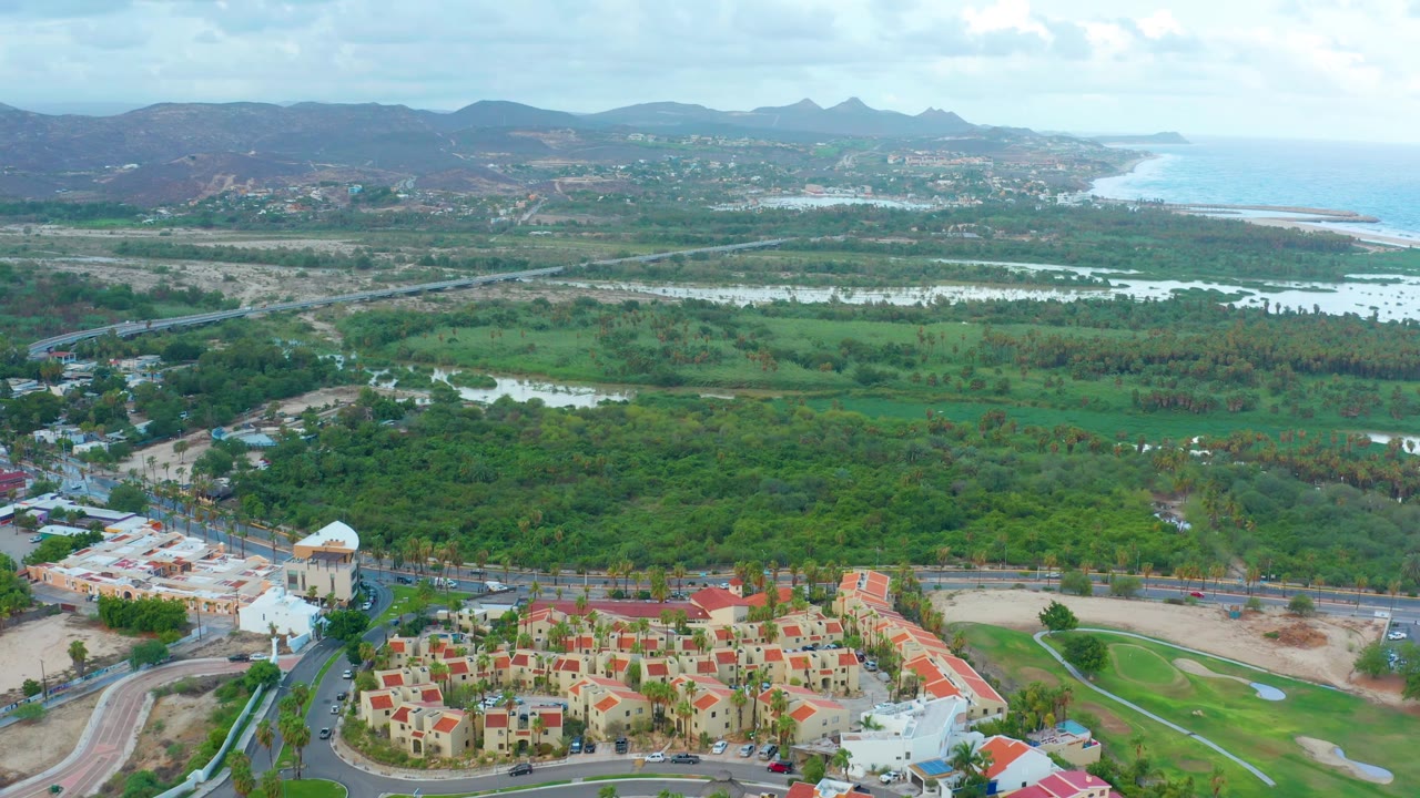 San Jose del Cabo aerial frame with a compact red-roofed housing area in the lower right, a white domed structure nearby, green fields and water channels through the middle, coastline at right, and no visible people.