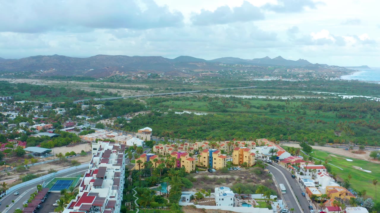 Drone view of residential blocks, curving roads, green vegetation, water channels, mountains, and coastline.
