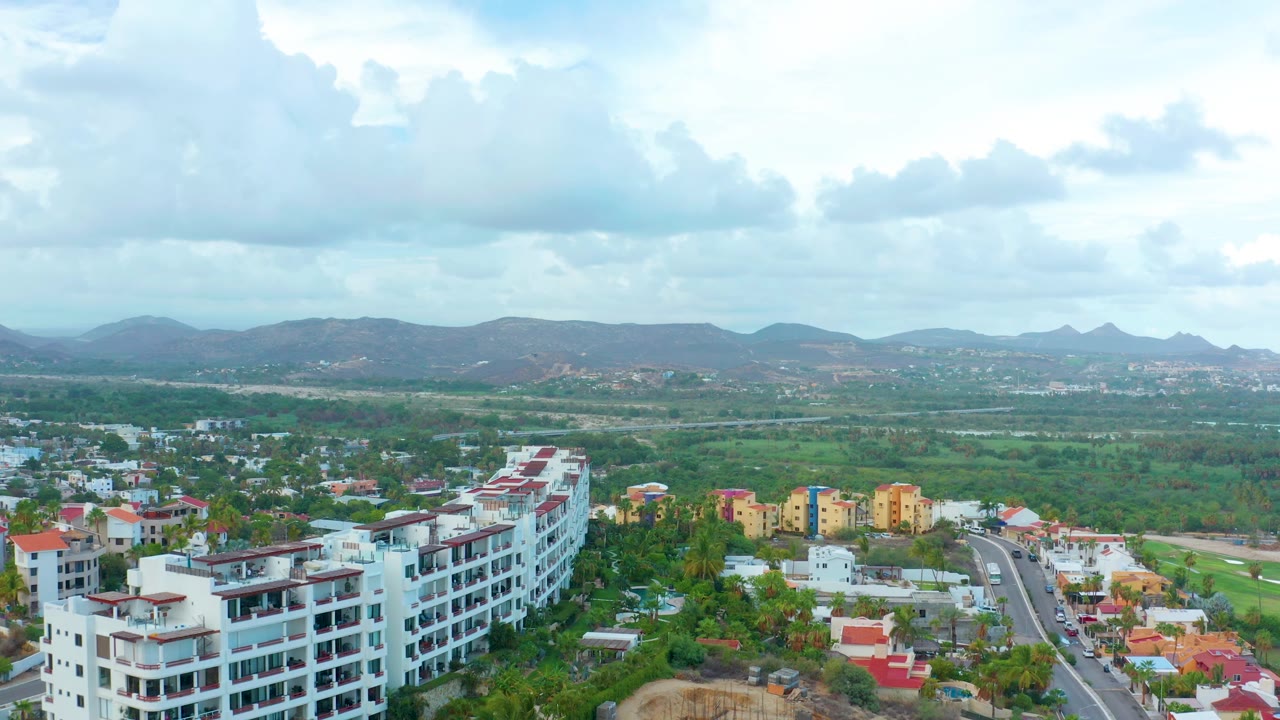 Aerial frame over San Jose Del Cabo showing white apartment buildings in the foreground, low-rise homes and greenery beyond, distant hills, and no visible people.