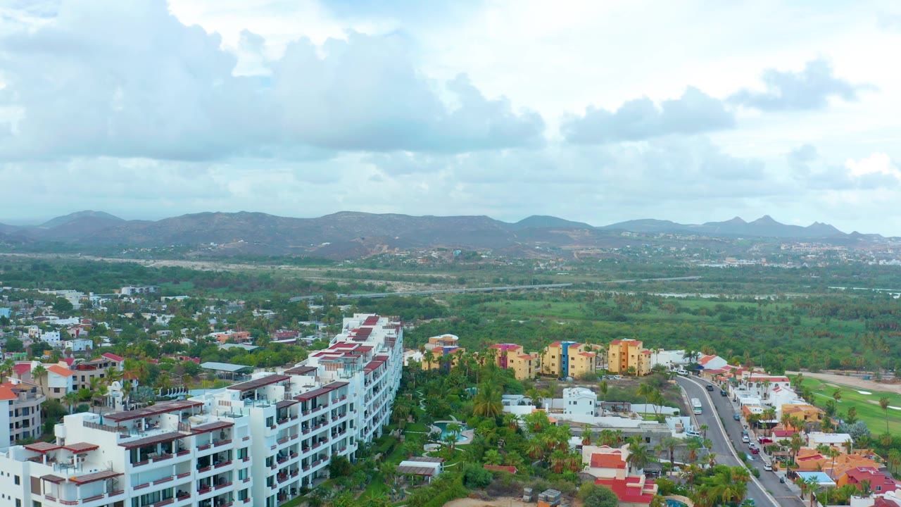 Aerial frame from San Jose Del Cabo with white and red-roofed buildings at the bottom, broad green midground, a low mountain range in back, and no visible people.