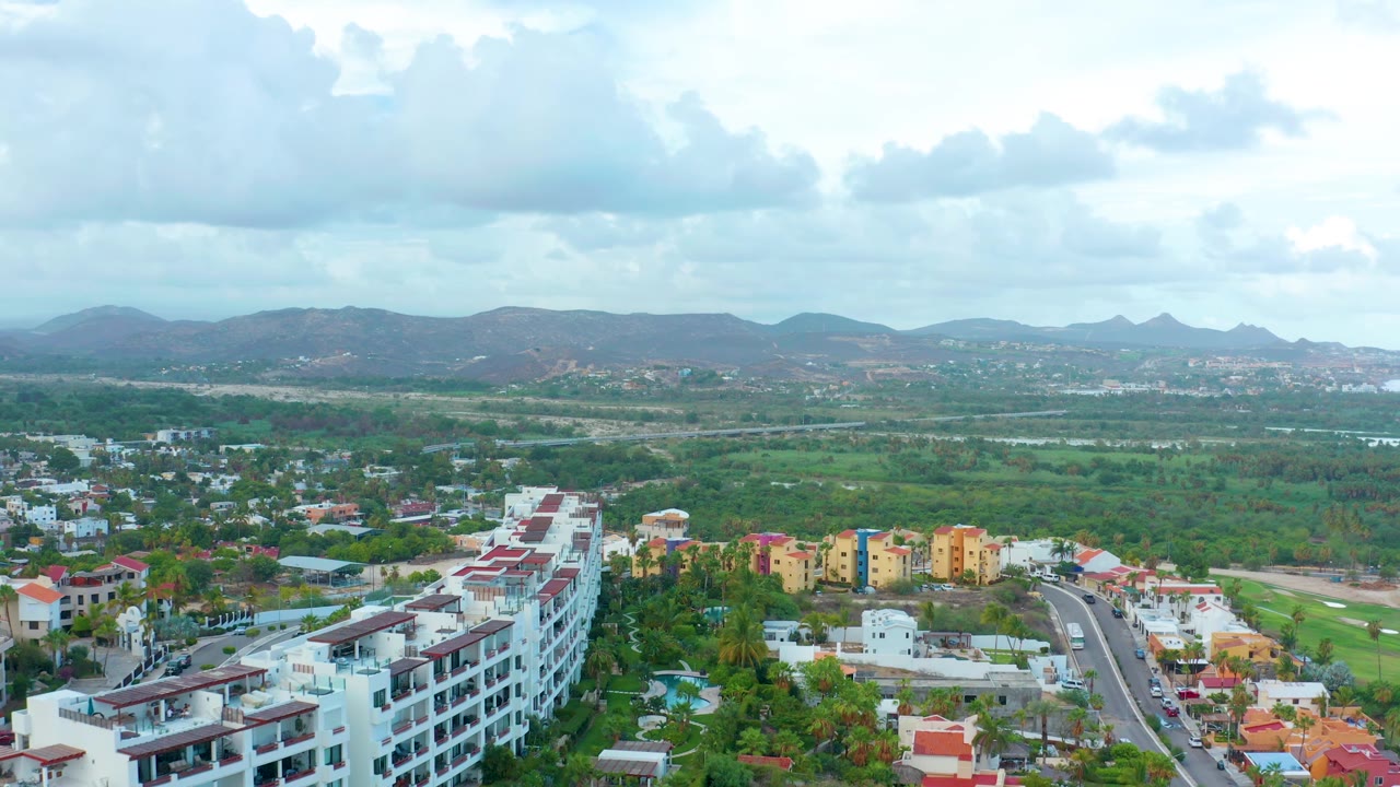 Aerial townscape with road, apartment rooftops, green terrain, and distant mountains.