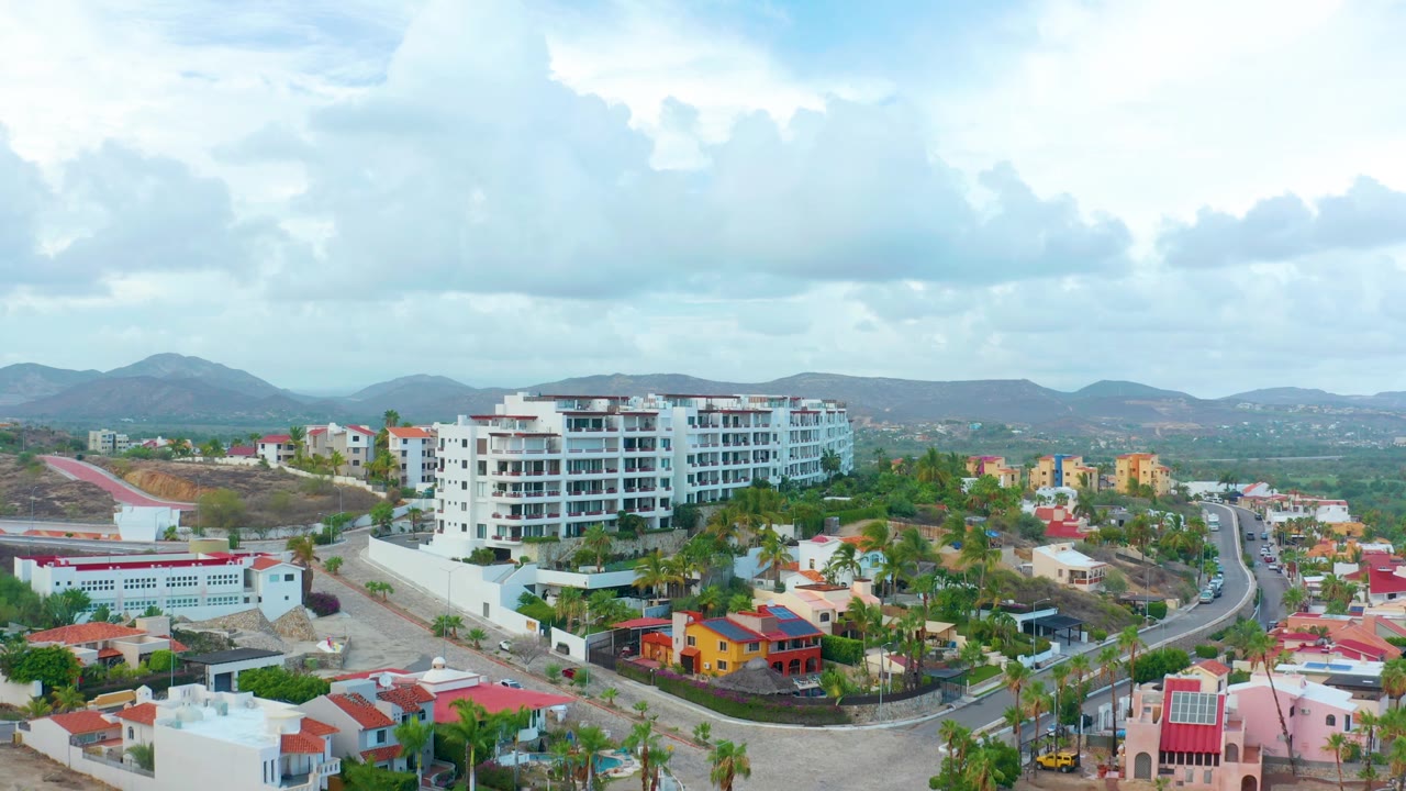 San Jose del Cabo aerial frame with a white apartment building left of center, red-roofed houses, palms, a curving road, distant hills, cloudy sky, and no visible people.