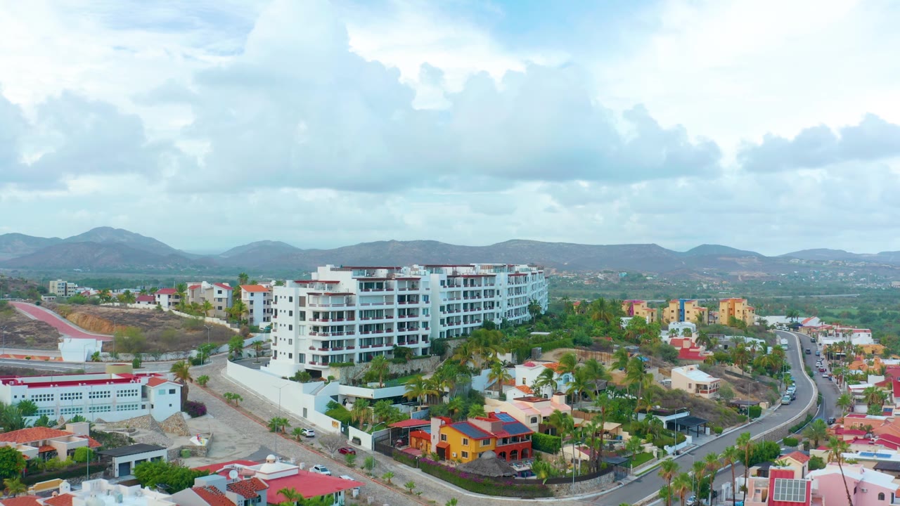 San Jose del Cabo aerial frame where the white apartment building shifts slightly right, a paved road bends through the neighborhood, low houses sit in the foreground, hills line the background, and no visible people appear.