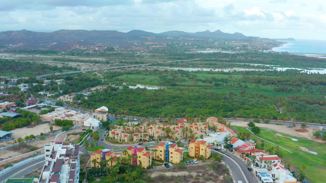 Opening aerial frame over San Jose Del Cabo with red-roofed residential blocks in the foreground, green fields and water beyond, distant mountains, coastline, overcast sky, and no visible people.