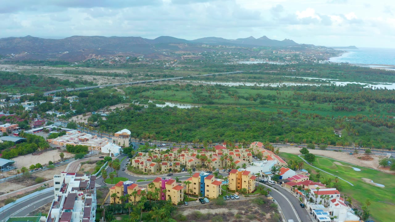 Drone view over red-roofed housing, roads, green fields, water, and distant hills.